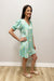 Woman wearing a light green patterned dress standing against a plain white wall.