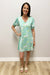Woman wearing a light blue patterned dress standing against a white wall.