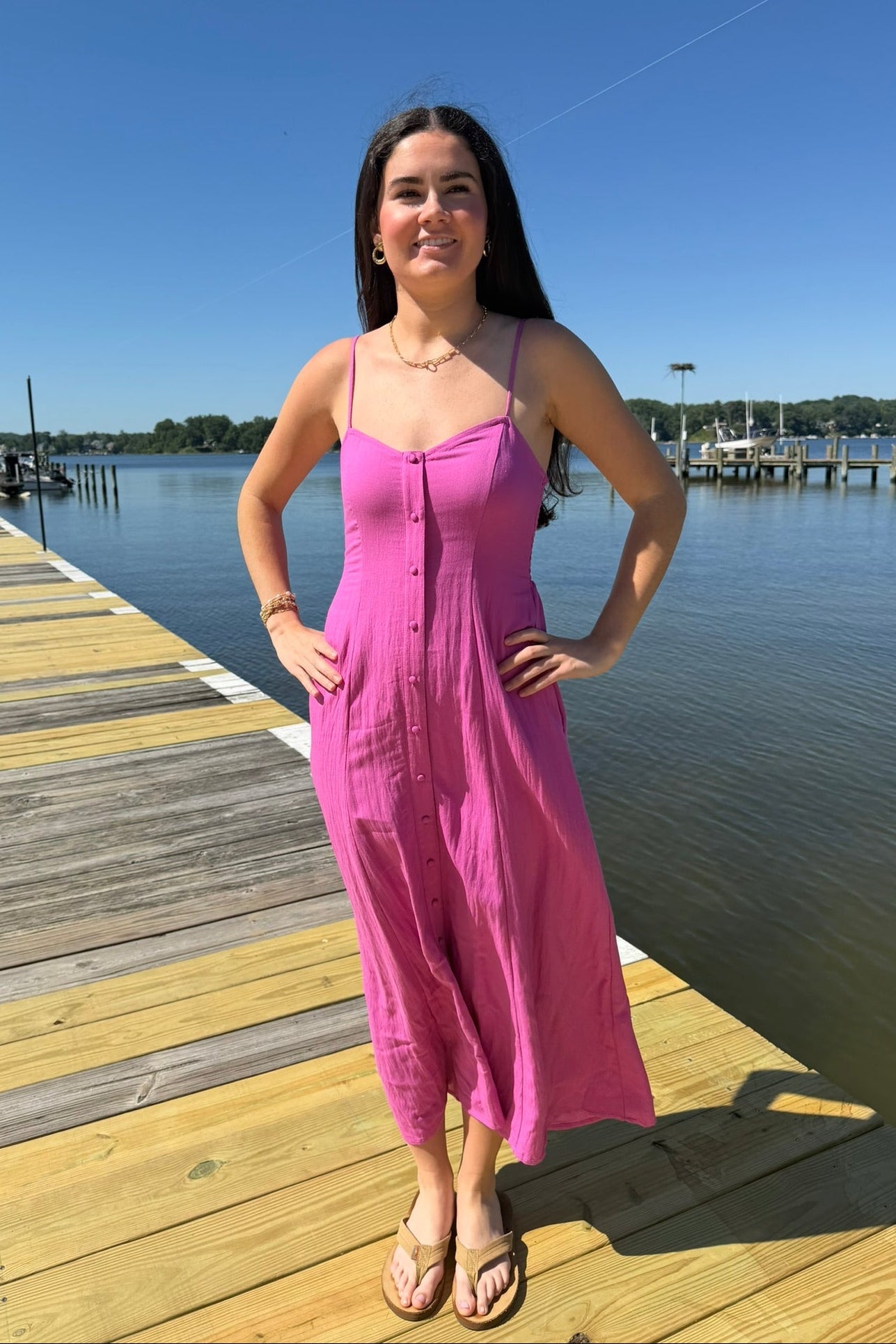 Woman in a pink dress standing on a wooden dock by a body of water.