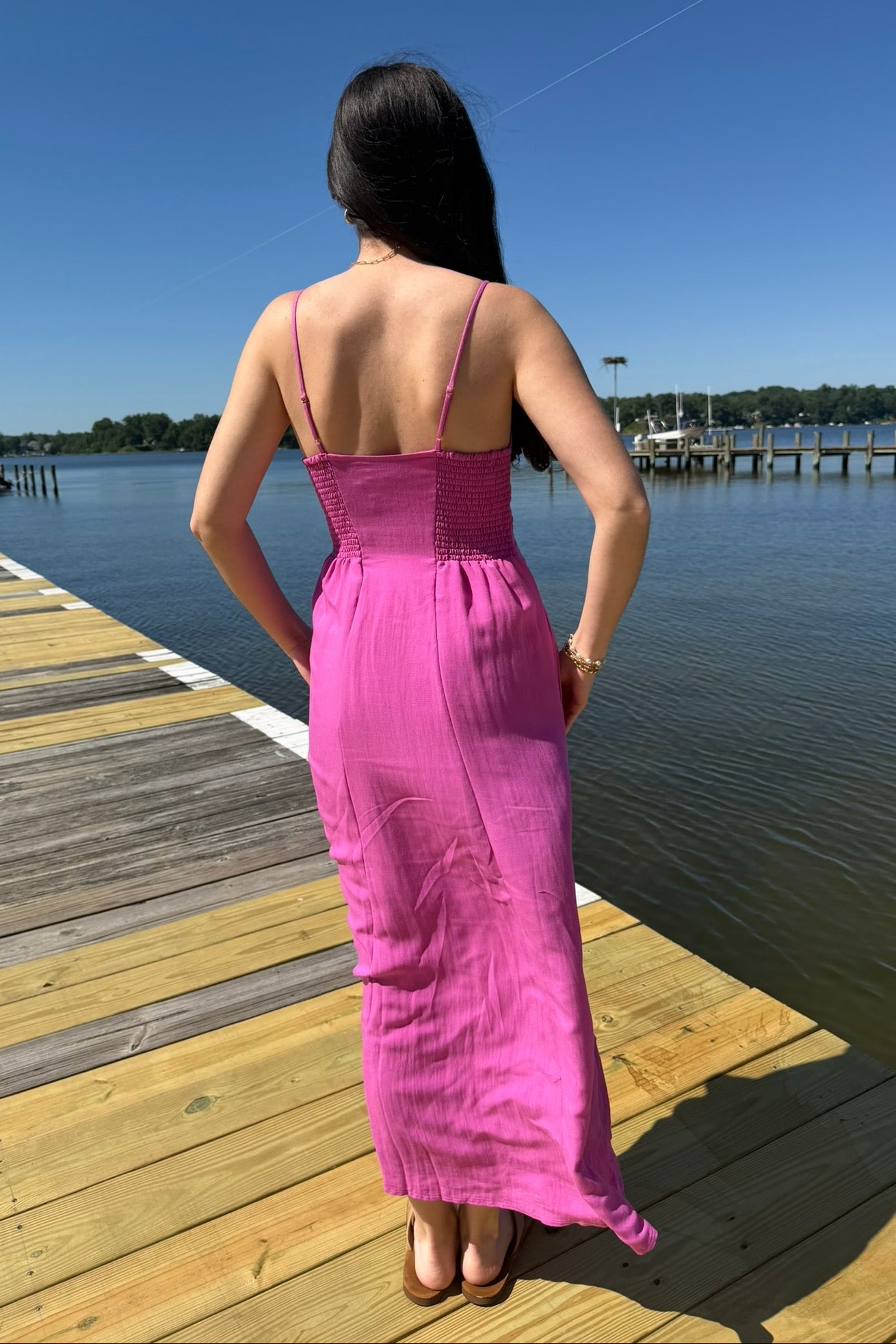 Woman in a pink dress standing on a wooden dock by a body of water.