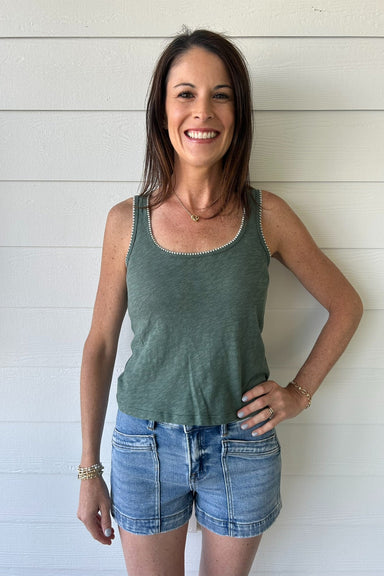 Woman wearing a green tank top and denim shorts standing against a white wooden wall.