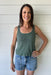 Woman wearing a green tank top and denim shorts standing against a white wooden wall.